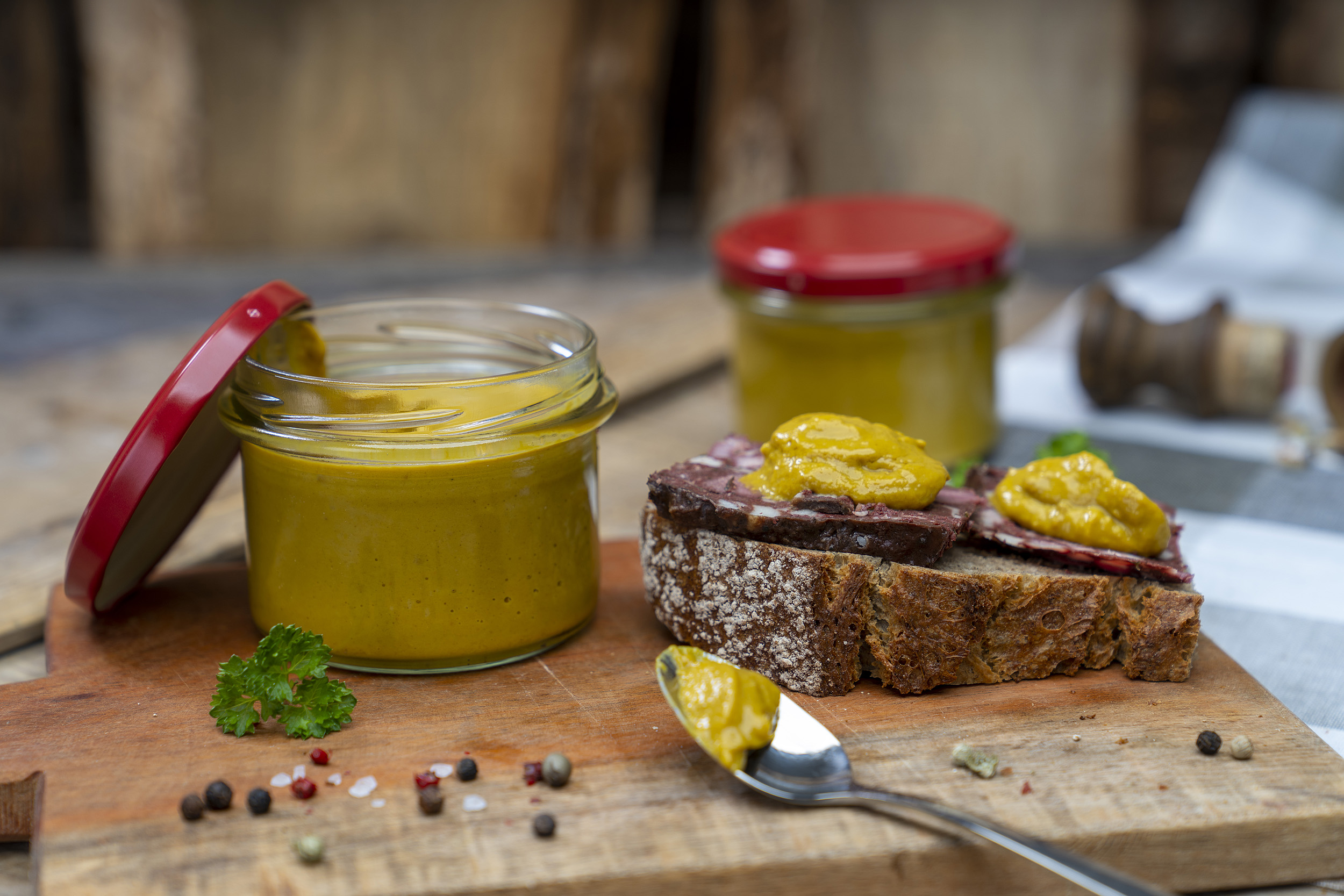 Meisters Mostrich Auf einem Holzbrett ein Glas Senf bzw. Meisters Mostrich. Daneben eine Schnitte mit Rotwurst und Senf. Darum Gewürze und Kräuter. Vorn ein Löffel mit etwas Senf. Hinten ein weiteres Glas.
