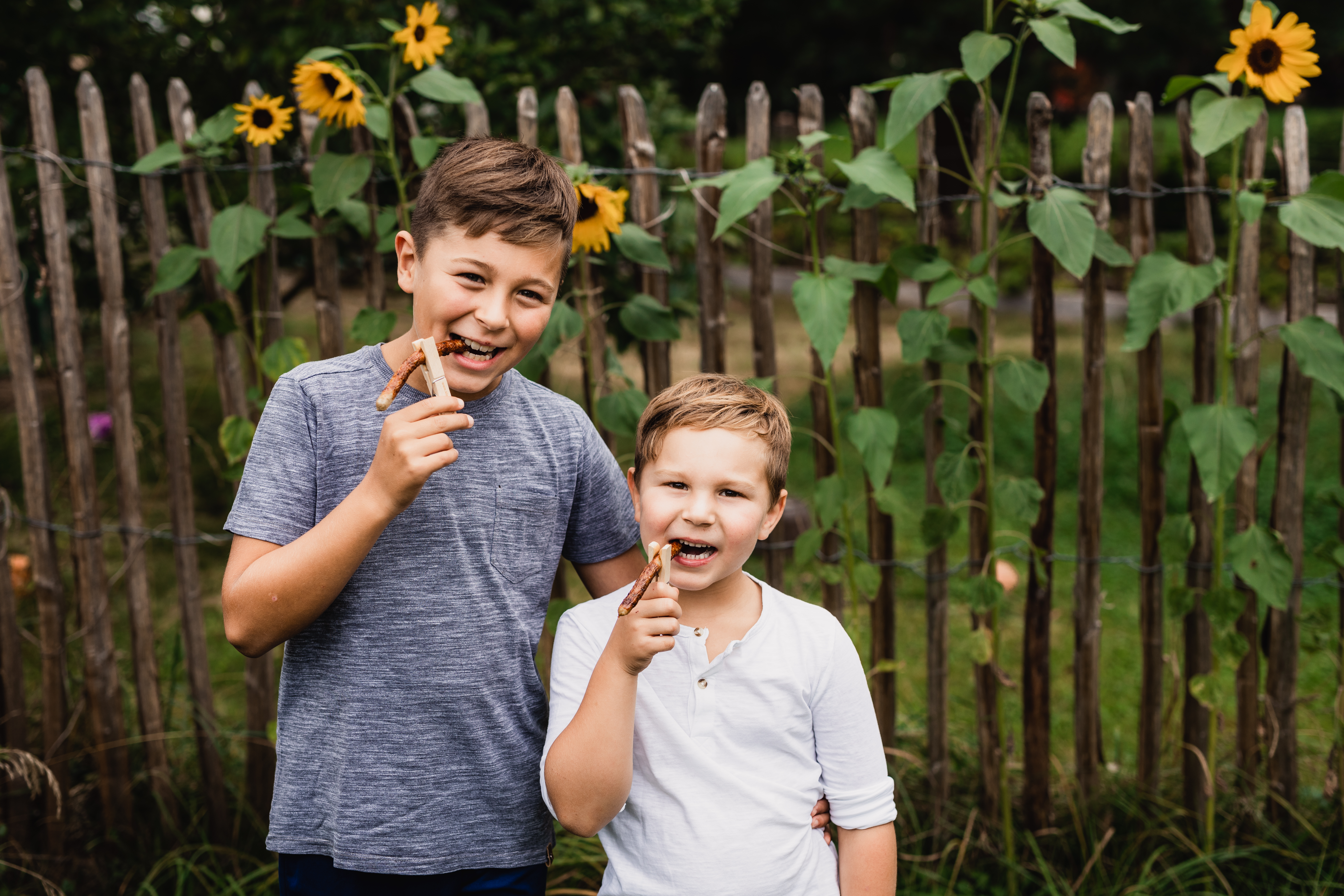 Zwei Jungen stehen nebeneinander und beißen von Klammerwürsten ab. Sie halten die Würste mit einer Holzklammer. Im Hintergrund ein Garten mit Sonennblumen.