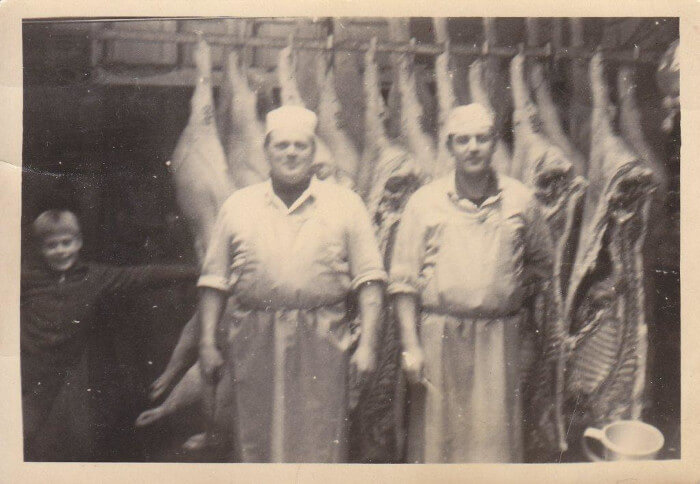 Gerhard und Bodo Fessel stehen vor zerteilten Schweinehälften. Links steht Erwin als kleiner Junge. Ein historisches Foto in schwarz-weiß.